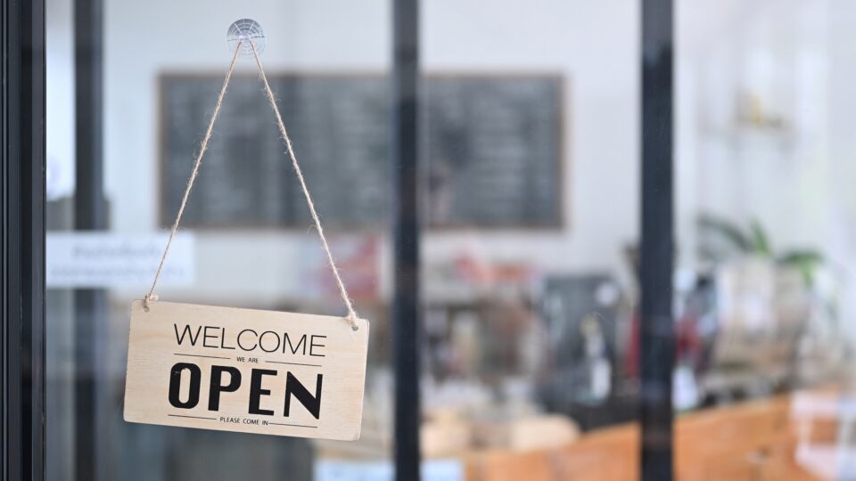 A "Welcome - Open" sign hangs in the window of a cafe for first steps to starting your own business blog.