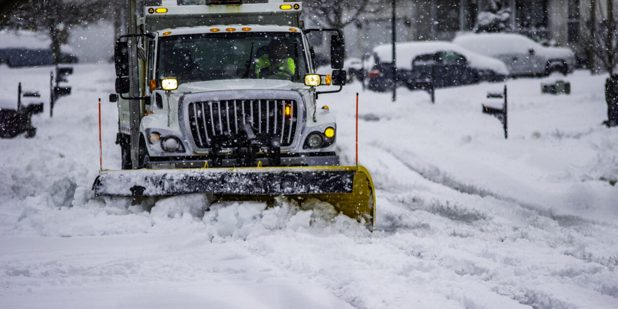 White snowplow service truck with orange lights and yellow plow blade clearing residential roads of snow while flakes are still falling why does a snow removal company need an answering service