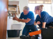 A male plumber fixes a leak in a bathroom sink while a female trainee observes, holding a digital tablet for the why-your-plumbing-business-needs-an-answering-service blog.