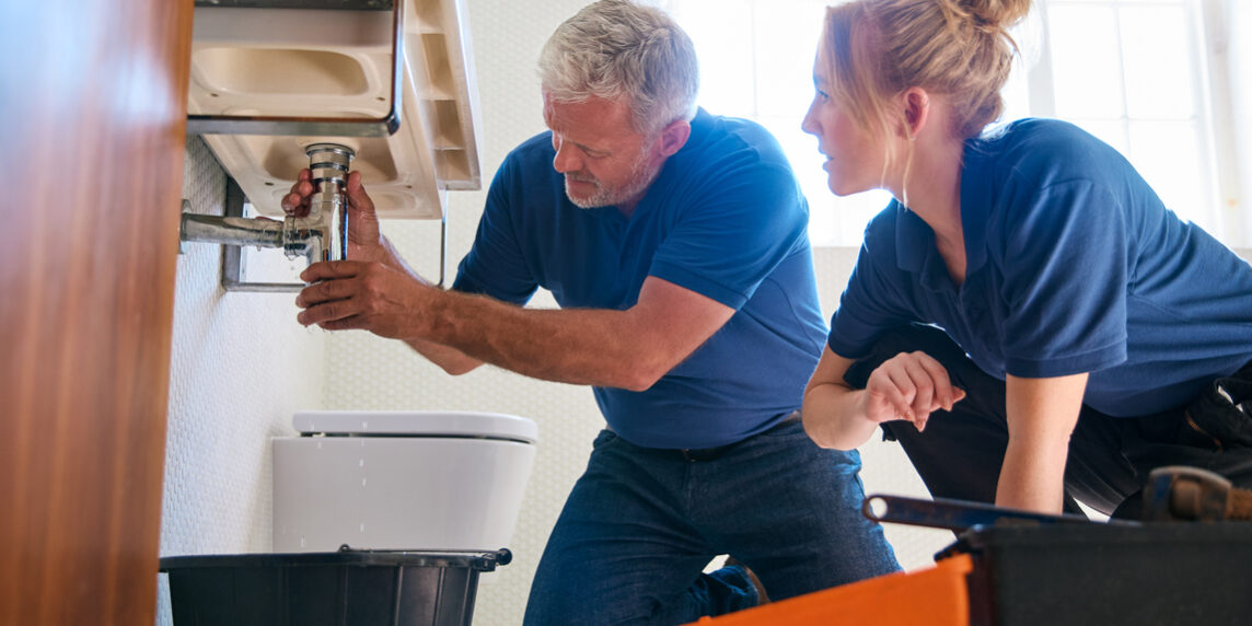Male Plumber Fixing Leak In Domestic Bathroom Sink With Female Trainee Holding Digital Tablet A male plumber fixes a leak in a bathroom sink while a female trainee observes, holding a digital tablet for the why-your-plumbing-business-needs-an-answering-service blog.