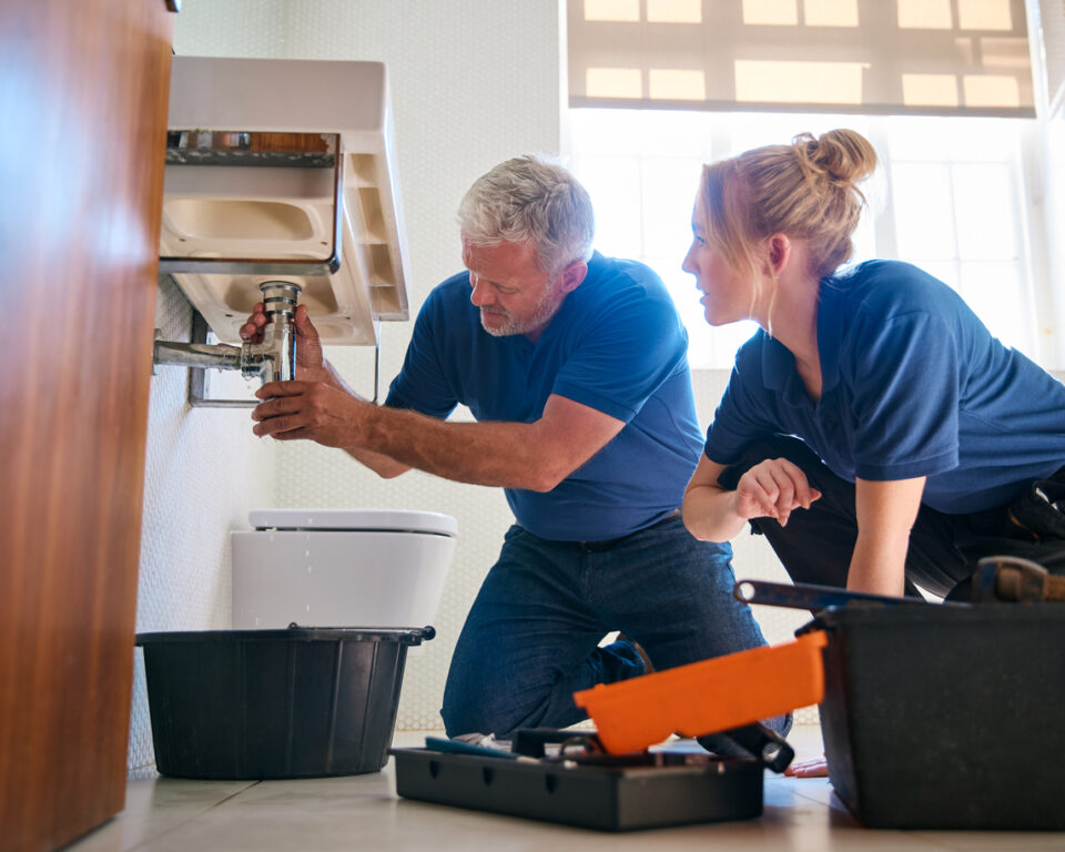 Male Plumber Fixing Leak In Domestic Bathroom Sink With Female Trainee Holding Digital Tablet A male plumber fixes a leak in a bathroom sink while a female trainee observes, holding a digital tablet for the why-your-plumbing-business-needs-an-answering-service blog.