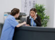 A receptionist and a medical assistant discuss paperwork at a check-in counter for urgent-care-answering-service blog.